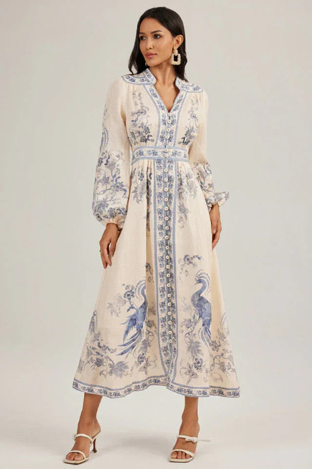Woman wearing a long, cream-colored maxi dress with a blue peacock print and long sleeves.