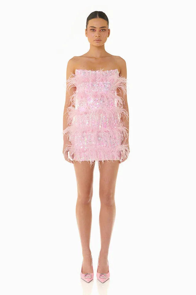 Model wearing a pink strapless sequin mini dress with feather trim, standing against a white background.