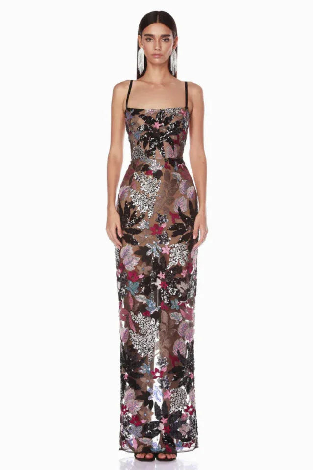Woman wearing a floor-length black sheer gown with colorful floral appliqués and sequins, featuring spaghetti straps and a straight neckline.