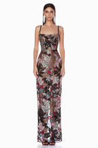 Woman wearing a floor-length black sheer gown with colorful floral appliqués and sequins, featuring spaghetti straps and a straight neckline.