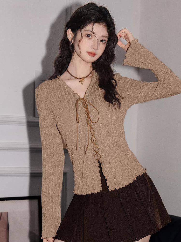Close-up of a woman wearing a tan ribbed knit long-sleeve top with a tie-front closure and ruffled hem, paired with a brown pleated skirt.