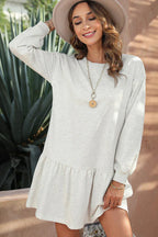 Woman wearing a light grey long-sleeved drop-waist dress with a ruffled hem and a cream fedora hat, accessorized with a gold pendant necklace and bracelets.