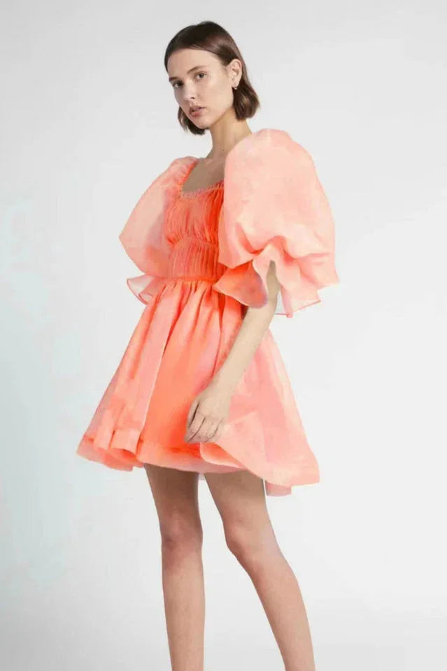 Woman wearing a short, coral organza dress with dramatic puff sleeves and a square neckline.