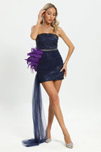 Woman wearing a navy blue strapless sequined mini dress with purple feather and navy blue tulle detailing on the hip and a flowing train, touching her hair, posing against a white background.