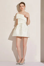 Full body shot of a woman in a white asymmetrical neckline dress with a short A-line skirt.