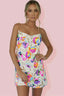 Woman wearing a white mini slip dress with colorful floral sequins and gold chain straps, posing against a pink background.