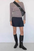Woman wearing a grey long-sleeved top, a black pleated mini skirt, and black knee-high boots with a black shoulder bag.
