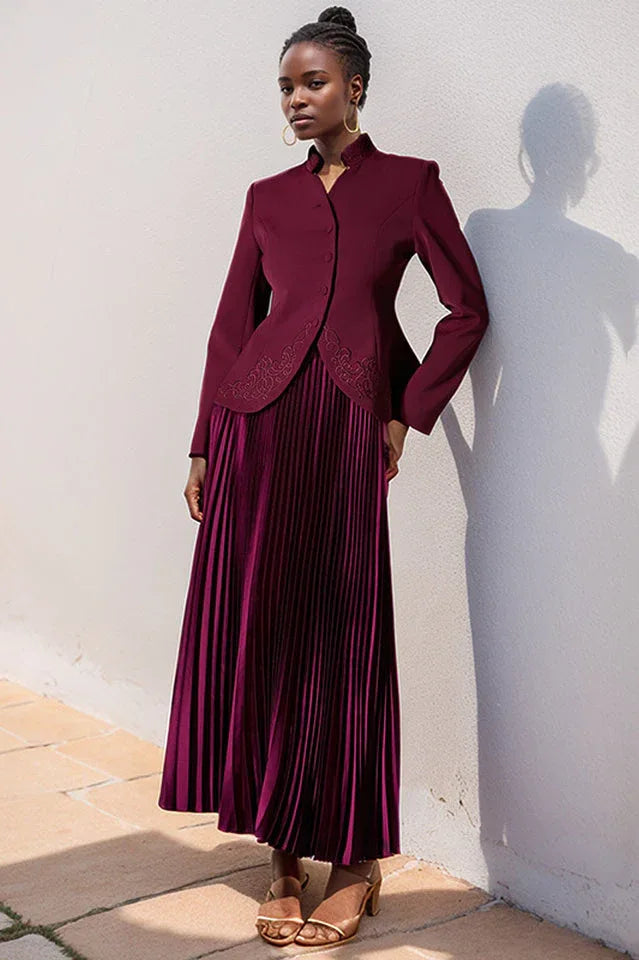 Woman wearing a burgundy jacket and pleated skirt, leaning against a white wall. Main image