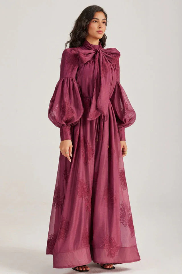 Woman wearing a long, deep purple dress with a pussy-bow neckline and bishop sleeves, featuring an embroidered floral pattern.