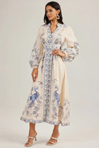 Model posing in a cream-colored, long-sleeved maxi dress featuring a prominent blue peacock pattern and a cinched waist.