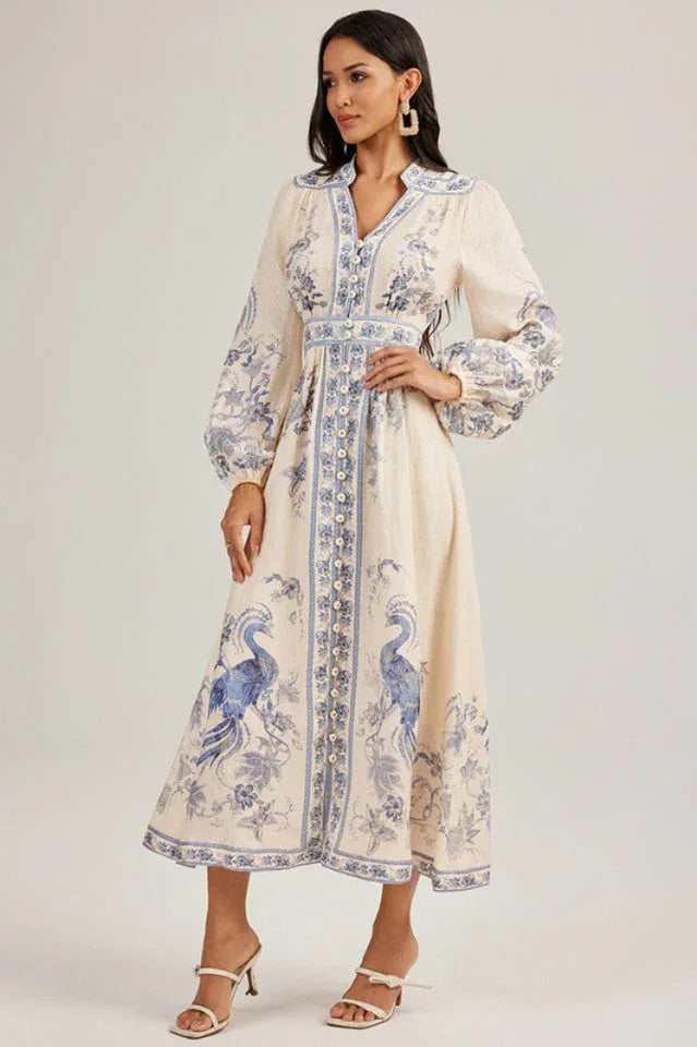 Model posing in a cream-colored, long-sleeved maxi dress featuring a prominent blue peacock pattern and a cinched waist.