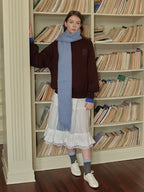 Woman wearing a brown velour sweatshirt with a light blue scarf, a white ruffled skirt, grey socks, and white sneakers, standing in front of a bookshelf.