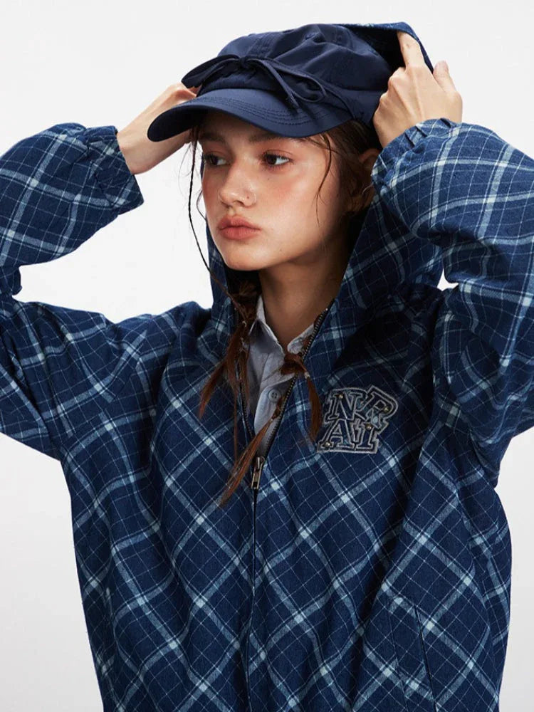 Model adjusting the hood of a blue plaid hooded jacket with an embroidered logo, wearing a light blue collared shirt and a navy blue cap.