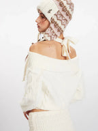 Back close-up of a model wearing a cream off-the-shoulder knit sweater and a matching mini skirt, with an ear flap hat and pink handbag.