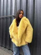Woman wearing a yellow faux fur jacket with blue jeans and beige boots, standing against a corrugated metal wall.