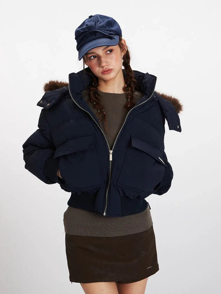 Front view of a woman wearing a navy cropped puffer jacket with a fur-trimmed hood, a brown knit top, and a navy baseball cap. She is wearing the jacket unzipped.