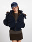 Front view of a woman wearing a navy cropped puffer jacket with a fur-trimmed hood, a brown knit top, and a navy baseball cap. She is wearing the jacket unzipped.