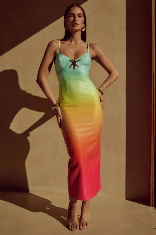 Full-length image of a model wearing a teal, yellow, orange, and pink ombre sequined midi dress with spaghetti straps and a dragonfly embellishment at the bust, standing against a tan wall with shadows.