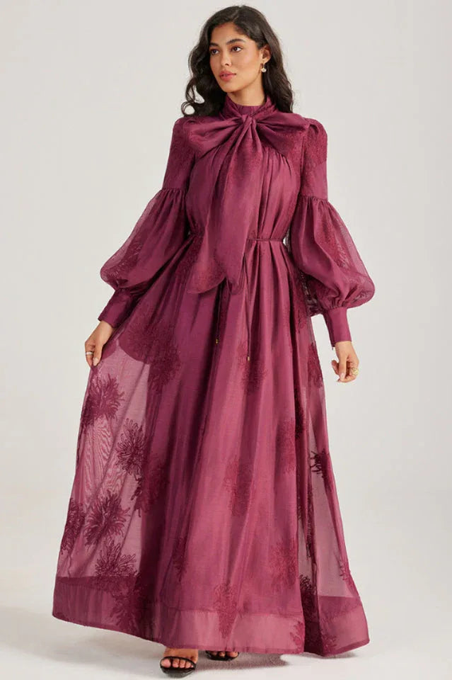 Close-up of a woman wearing a long, deep purple dress with a prominent pussy-bow neckline and embroidered details.