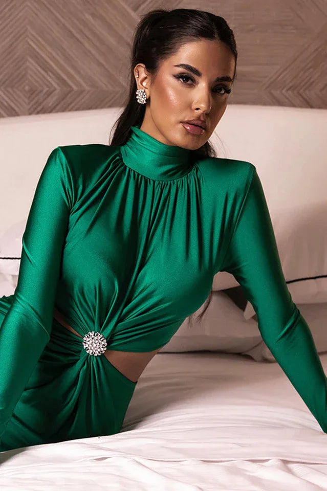 Woman wearing a long-sleeved green satin dress with a cut-out waist and embellished detail, sitting on a bed, looking forward.