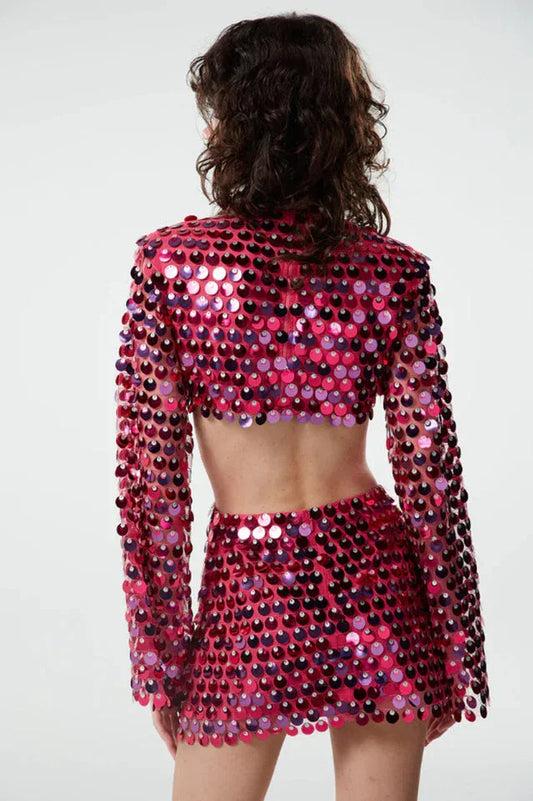 Back view of a woman wearing a pink sequined crop top with bell sleeves and a matching mini skirt with a cut-out detail at the waist.