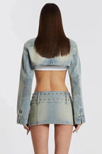 Back view of a woman wearing a light wash denim cropped jacket and a matching mini skirt with buckle detailing.