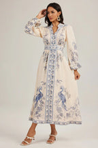 Woman touching her hair while wearing a cream-colored maxi dress with a blue peacock print and puffed sleeves.