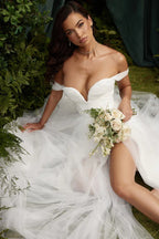 Model sitting on the ground in a white off-the-shoulder wedding dress with a high slit, holding a bouquet.