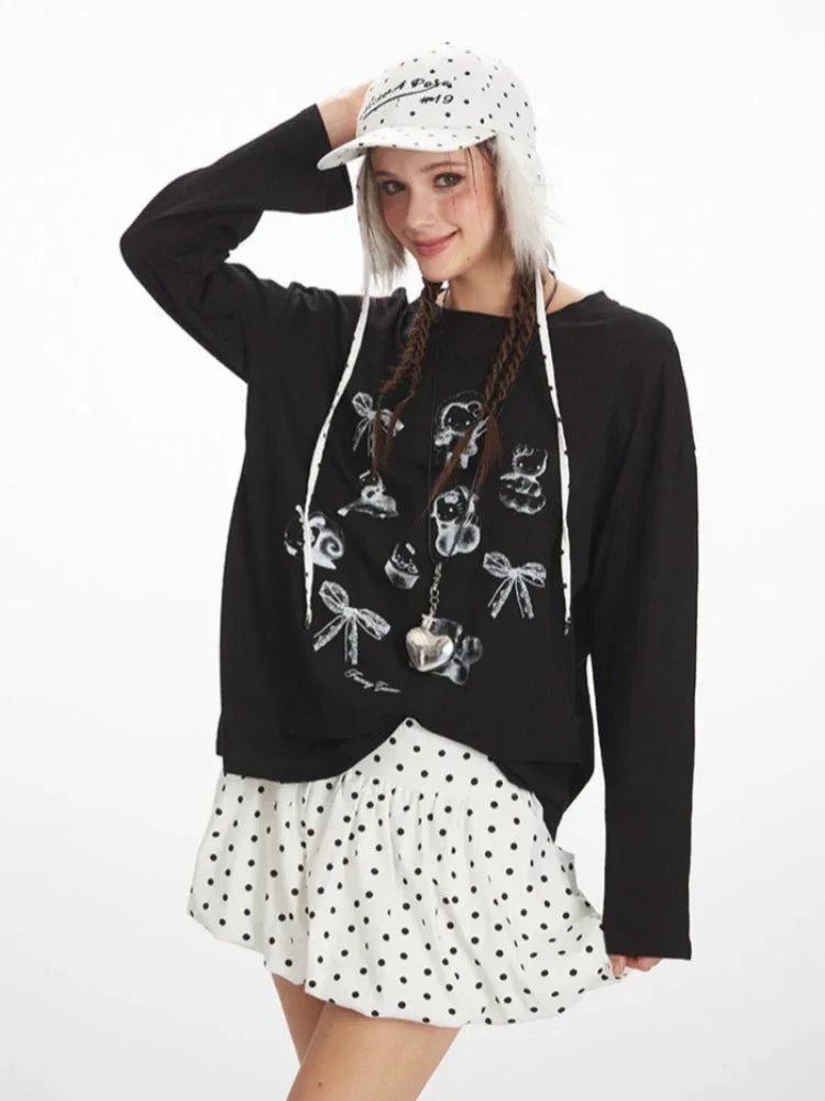 Medium shot of a young woman wearing a black long-sleeved graphic t-shirt and a white polka dot mini skirt. She is smiling and touching her hat, which has fur trim.