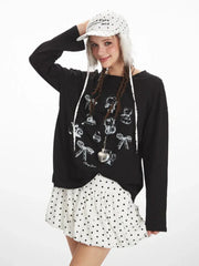 Medium shot of a young woman wearing a black long-sleeved graphic t-shirt and a white polka dot mini skirt. She is smiling and touching her hat, which has fur trim.