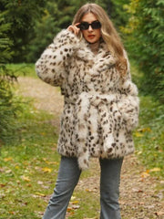 Woman wearing a spotted faux fur coat cinched with a belt, grey jeans, and sunglasses, standing outdoors on a path.