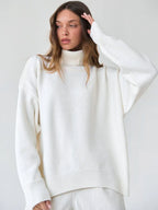 Woman with her hand on her head, wearing a white turtleneck sweater and white pants.