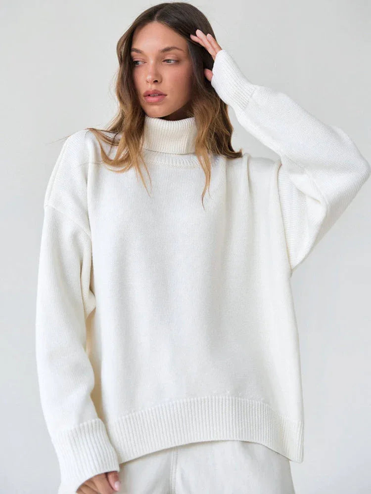 Woman with her hand on her head, wearing a white turtleneck sweater and white pants.