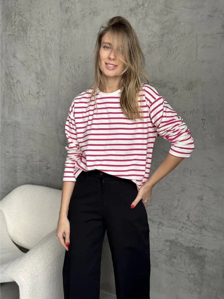 Woman wearing a white and red striped long-sleeve top and black pants, posing against a grey wall.