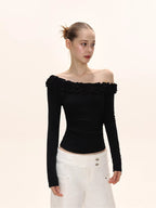 Side view of a model in a black off-the-shoulder long-sleeved top with ruffle detailing, and white wide-leg pants.