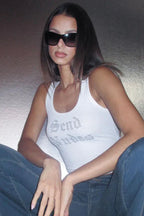 Woman wearing a white ribbed knit tank top with rhinestone "Send Nudes" graphic and blue jeans, sitting down