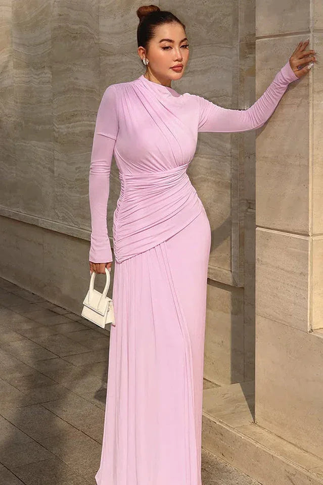 Woman in a long pale pink ruched dress holding a white handbag.