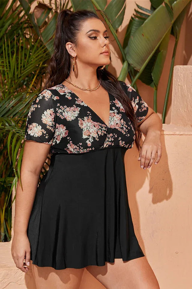 Plus-size woman in a black floral print one-piece swimsuit with short sleeves and a skirted bottom, posing outdoors.