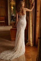 Back view of a woman in a white, beaded mermaid wedding dress with a high slit and train, leaning against a curtain.