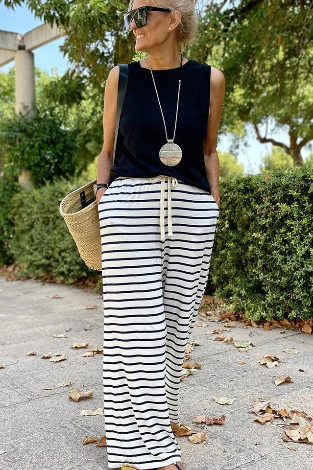 Woman from the front wearing a black sleeveless top and black and white striped wide leg pants, holding a straw tote bag.