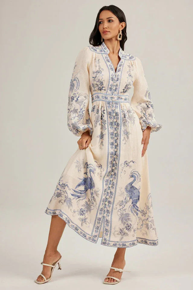 Woman adjusting the hem of a flowing, cream-colored maxi dress adorned with a blue peacock print.