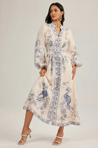 Woman adjusting the hem of a flowing, cream-colored maxi dress adorned with a blue peacock print.