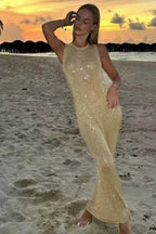 Woman wearing a gold sleeveless floor-length gown with sparkle embellishments, standing on a beach at sunset.