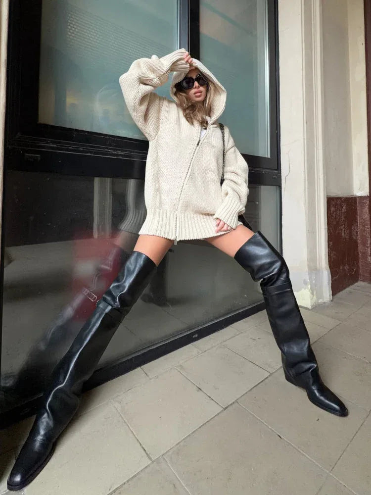 Woman wearing a beige oversized knit hooded sweater dress and black thigh-high boots, in a wide stance.