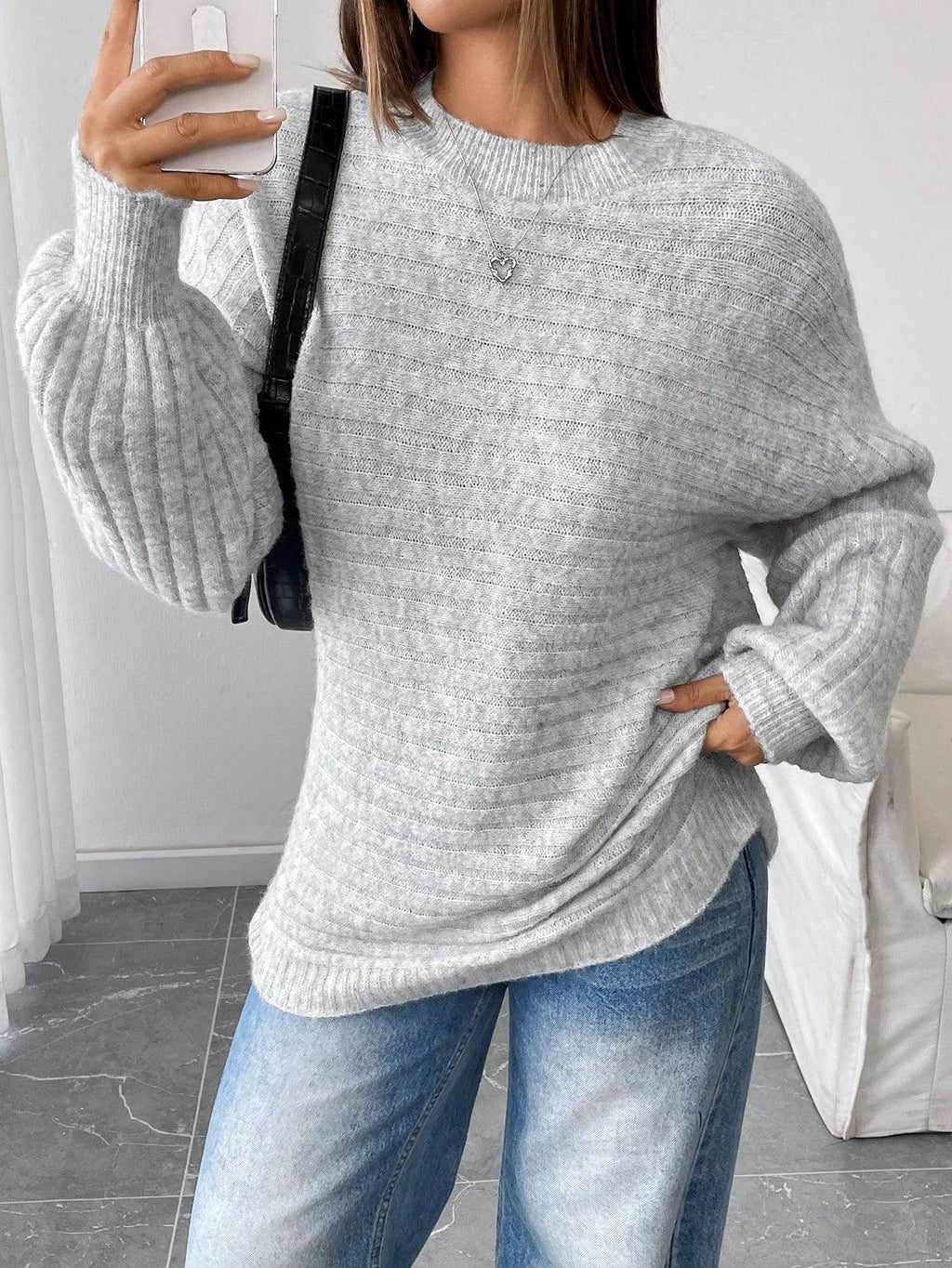 Woman taking a selfie while wearing a light grey oversized knit sweater with balloon sleeves and a crew neck, paired with blue denim jeans and a black handbag.
