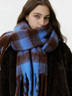 A woman's face is partially obscured by a large, oversized blue and brown plaid scarf with fringe.