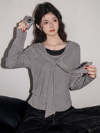 Woman wearing a grey long-sleeved top with a draped overlay and black pants, holding a small silver camera.