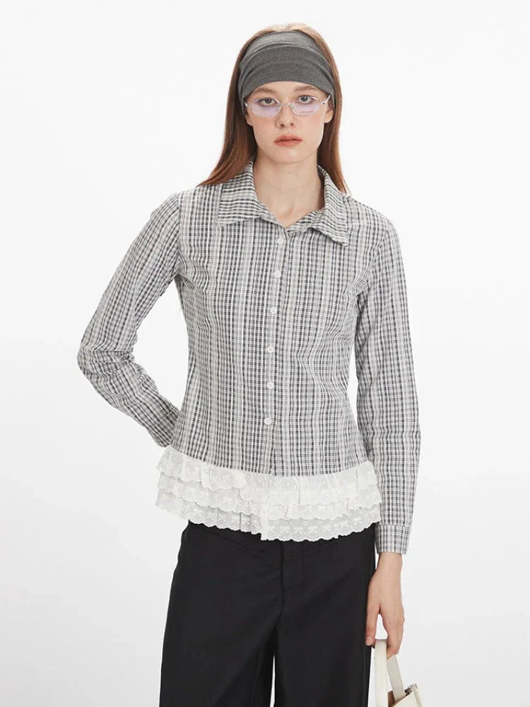 Front close-up of a woman wearing a long-sleeved plaid collared shirt with a white ruffled hem and black pants, with hands on hips.