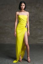 Woman in a yellow strapless gown with a high slit and floral embellishment standing against a concrete wall.