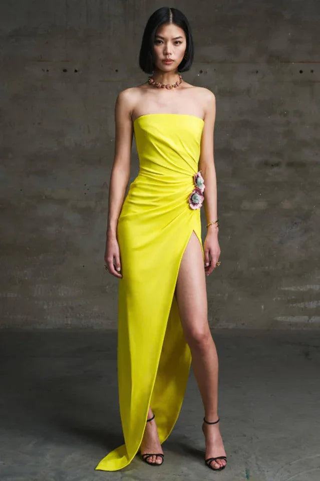 Woman in a yellow strapless gown with a high slit and floral embellishment standing against a concrete wall.